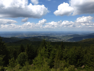 Das Foto zeigt eine Landschaft in der Region Südbaden. Aufgenommen von einem Berg aus. Im Vordergrund ein Wald. Lässt man den Blick weiter über den Wald weg schweifen folgen Hügel, teils besetzt mit Häusern einer Ortschaft. Bis zur Bildmitte erstreckt sich an die Hügel eine Ebene. Vereinzelt sind Häuser von Ortschaften und Wälder zu erkennen. Ab Bildmitte aufwärts ist ein teils mit weißen Wolken verhangener hellblauer Himmel zu sehen.