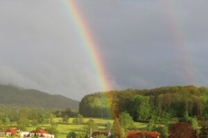 Landschaftsfoto mit einem Regenbogen in der Mitte des Bildes der sich von oben bis unten durchzieht. Im Hintergrund zu circa zwei Dritteln nach oben ein dunkel wolkenverhangener Himmel. Im unteren Drittel eine Wiese mit Bäumen und zwei Wälder.